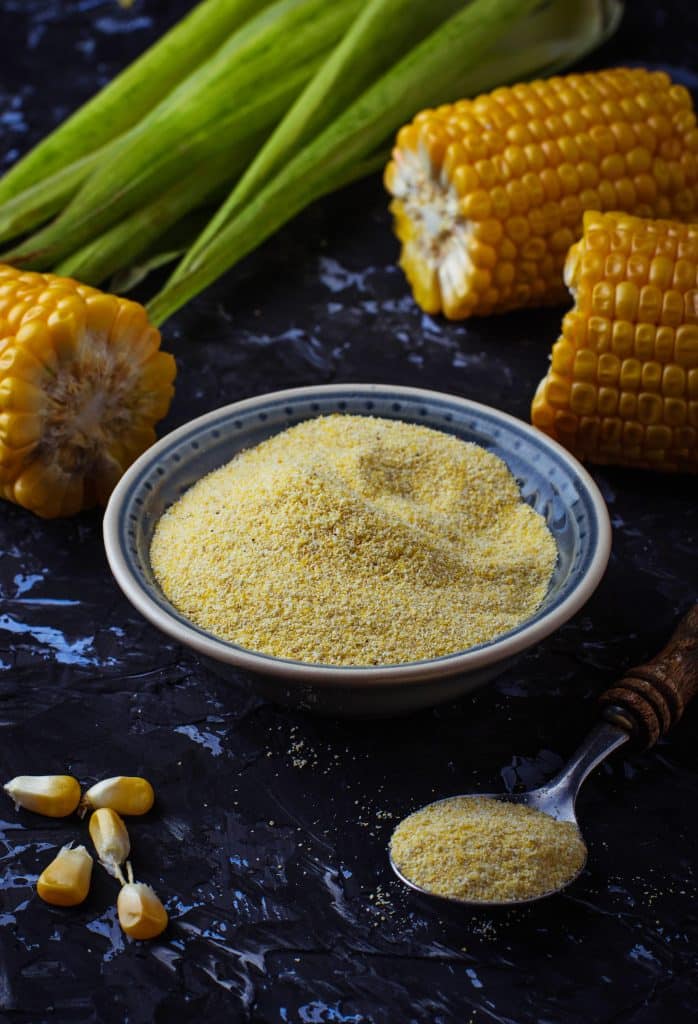 Cornmeal in a bowl with a spoon and corn on the cob