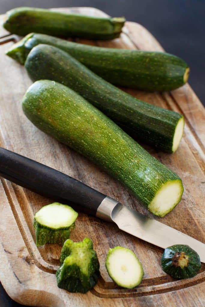 A whole zucchini next to a knife on a wooden board